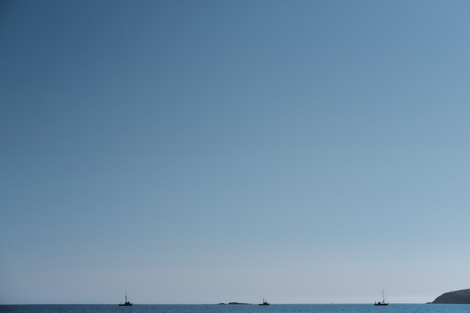 Wide blue sky over a calm sea, with three small boats and a headland on the horizon